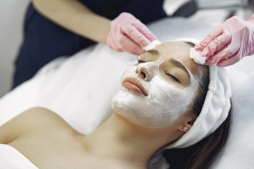 From above anonymous medical specialist in uniform and gloves wiping cream mask off face of relaxed woman during skincare procedure in modern studio